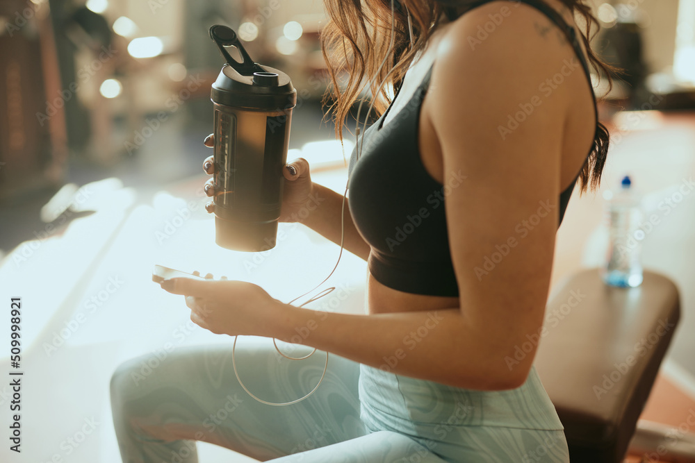 Cropped picture of a strong sportswoman sits in a gym, drinks protein drink and using a phone while drinking protein drink and taking a break. - obrazy, fototapety, plakaty 