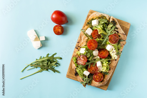 Pinsa with cheese, tomatoes, cabbage and arugula and the ingredients from which it is made on a light blue background.