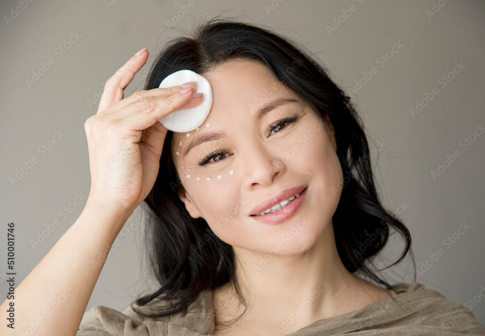 Portrait of beautiful Asian woman smiling and cleaning her face with cotton pads covered in cream enjoying beauty treatments in SPA salon Portrait of beautiful Asian woman smiling and cleaning her face with cotton pads covered in cream enjoying beauty treatments in SPA salon