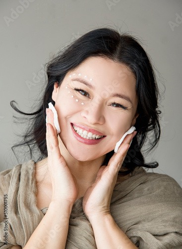 Portrait of beautiful Asian woman smiling and cleaning her face with cotton pads covered in cream enjoying beauty treatments in SPA salon