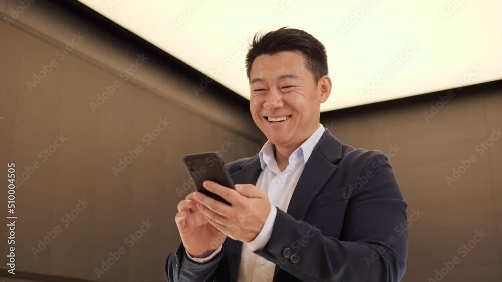 Confident korean man using mobile phone app in modern light office. Positive asian business guy browsing financial news smiling reading good news standing indoor. Successful male chinese adult. Confident korean man using mobile phone app in modern light office. Positive asian business guy browsing financial news smiling reading good news standing indoor. Successful male chinese adult.