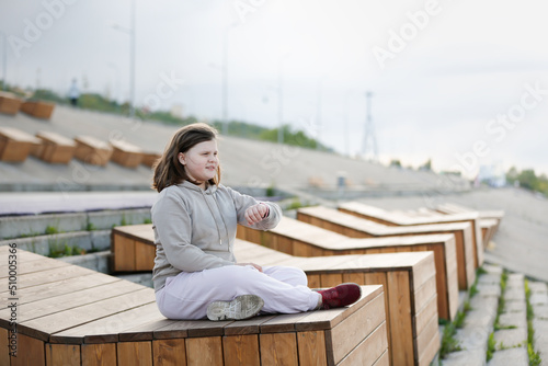 cute overweight teenager girl in tracksuit meditates, rests on wooden seat on concrete embankment