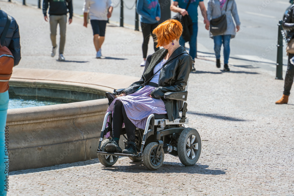 A disabled person in a wheelchair on a sunny day on a city street among healthy people looks at the fountain. Integration of people with disabilities into society