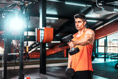 Shot of a muscular guy in sportswear working out with dumbbells at the cross training gym. He is pumping up shoulders muscule with heavy weight..