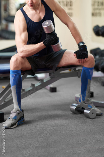 Cropped shot of an athletic man in a sportswear and blue knee socks with a color print. He is sitting down on a bench with a dumbbell in his hand. The man in golfs is isolated on the gym background.