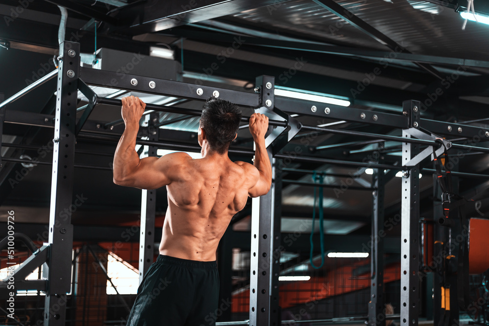 Fototapeta premium Back view of muscular sportsman doing pull ups on horizontal bar at gym. Sports trainer in gym workout exercising. Fit and athletic man having naked torso, wearing in black shorts..