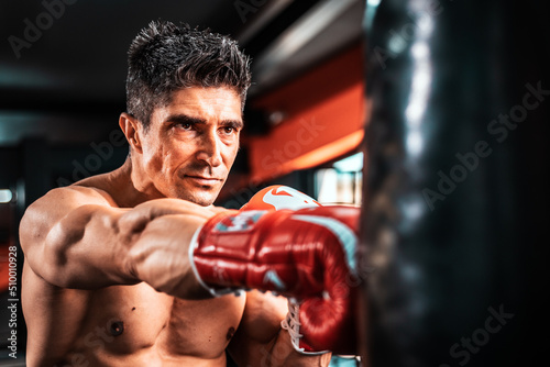 Caucasian fighter man wear boxing red gloves and punching forward with one fist to camera in the fitness sport gym. Make looking forward with serious face. Exercise for a healthy body. © Djordje