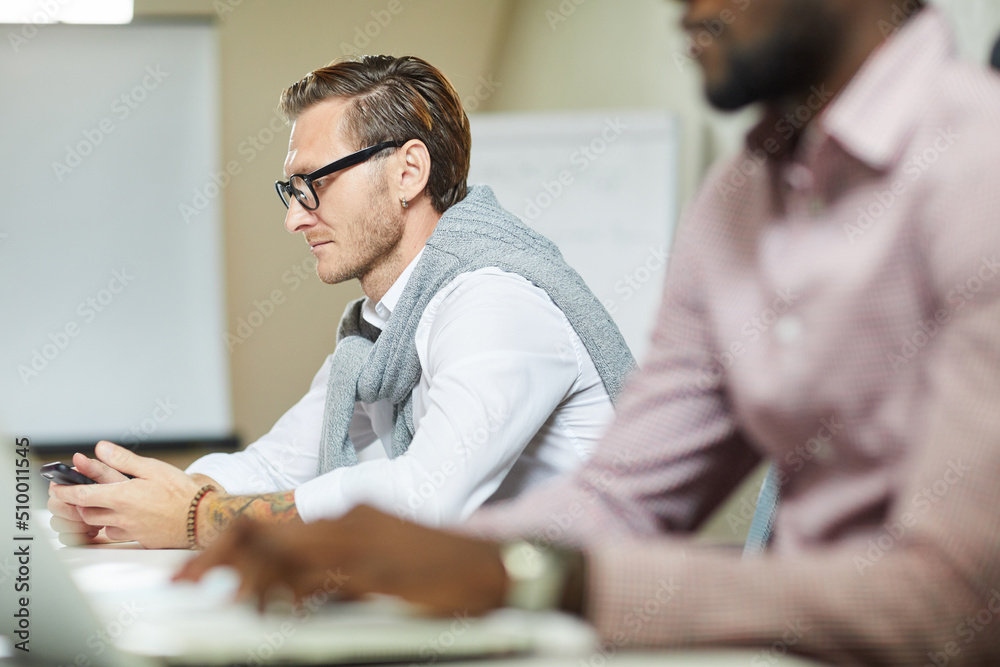 Serious young man in eyeglasses sitting at table and holding gadget while thinking about project development Serious young man in eyeglasses sitting at table and holding gadget while thinking about project development