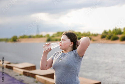 Overweight European teenage girl in tracksuit drinks water from bottle while jogging along on concrete embankment, Sports and overweight teenagers