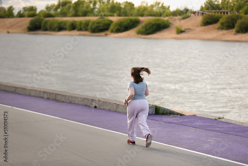 Leinwand Poster overweight European teenage girl in tracksuit jogging along concrete embankment on violet floor, outskirts of city