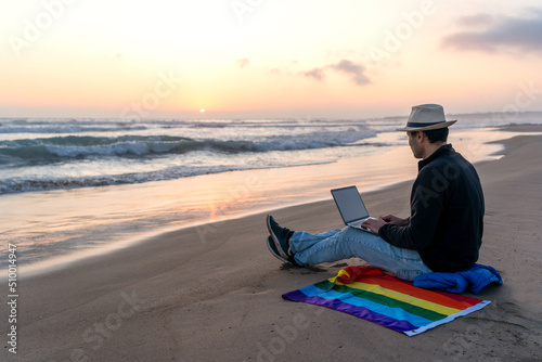 person on the shore of the beach with a laptop and a rainbow flag looking at the sunset on the horizon, LGTBQ
