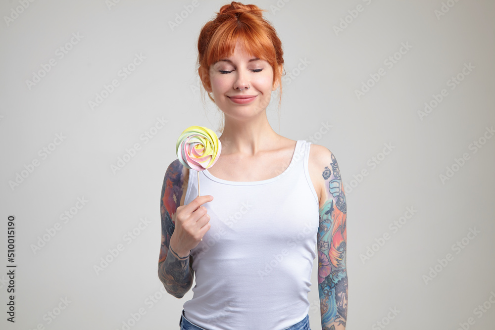 Indoor portrait of young ginger female posing over white wall holding big colorful candy and smiles Indoor portrait of young ginger female posing over white wall holding big colorful candy and smiles