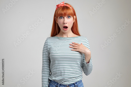 Indoor portrait of young ginger female posing over white wall looking into camera with surprised facial expression