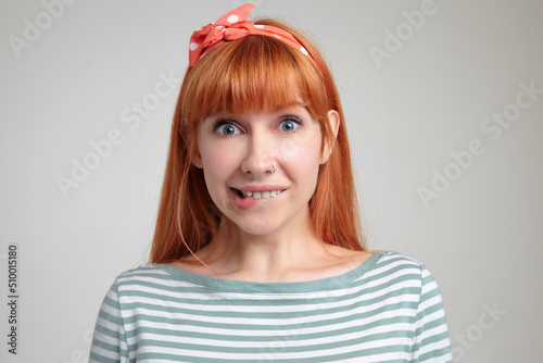 Indoor portrait of young ginger female posing over white wall looking into camera, biting her lip with excited facial expression
