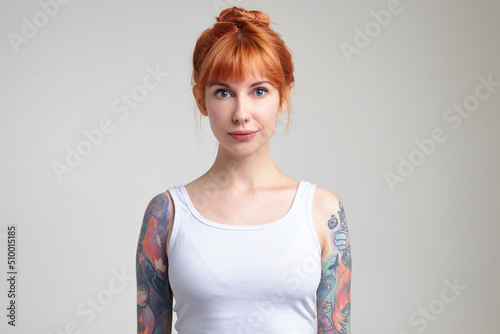 Indoor portrait of young ginger female posing over white wall looking into camera with calm facial expression