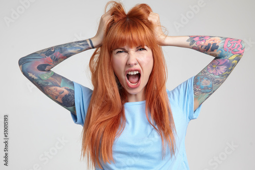 Indoor portrait of young ginger female posing over white wall looking into camera with angry facial expression