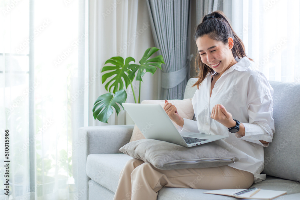 Portrait of a cheerful young asian girl sitting on sofa at home with laptop computer and celebrating success, smiling broadly looking happy, positive, confident with both arms up. Portrait of a cheerful young asian girl sitting on sofa at home with laptop computer and celebrating success, smiling broadly looking happy, positive, confident with both arms up.