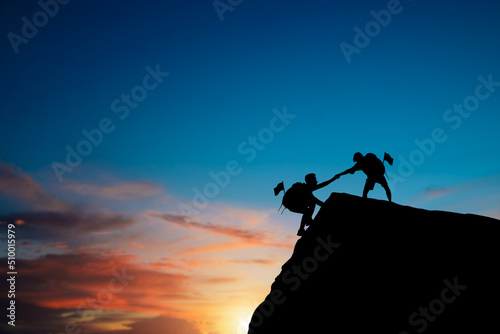 Silhouette of two people helping each other hike up on a mountain at sunrise. Giving a helping hand, and active fit lifestyle concept. Couple hiking help each other. Together they will succeed.