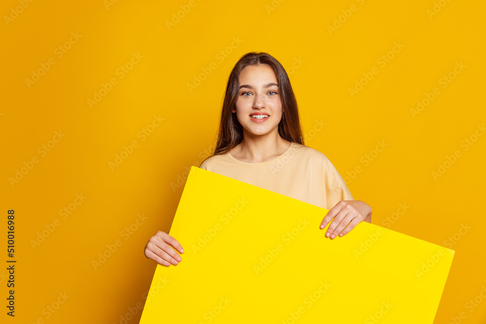 Young pretty girl, cheerful student or shop seller holding huge empty sheet of paper isolated on yellow background. Concept of beauty, art, fashion, youth and emotions Young pretty girl, cheerful student or shop seller holding huge empty sheet of paper isolated on yellow background. Concept of beauty, art, fashion, youth and emotions