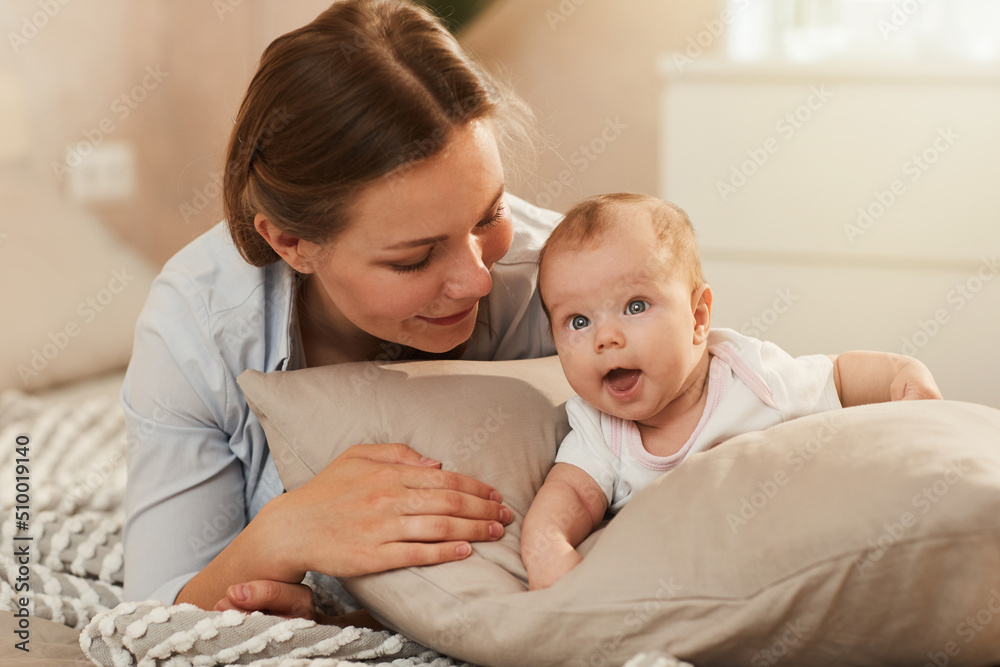 Smiling young mother looking at little happy baby crawling on soft pillow in bedroom