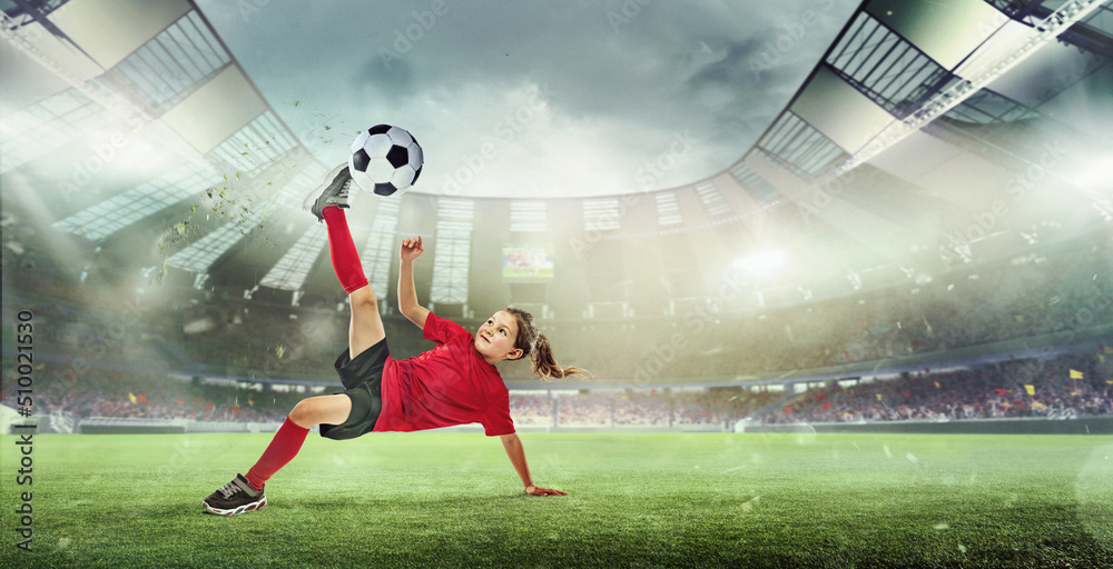 Portrait of little girl, football player in red uniform hitting ball and falling isolated over open air stadium.