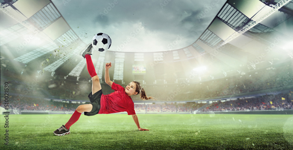 Naklejka premium Portrait of little girl, football player in red uniform hitting ball and falling isolated over open air stadium.