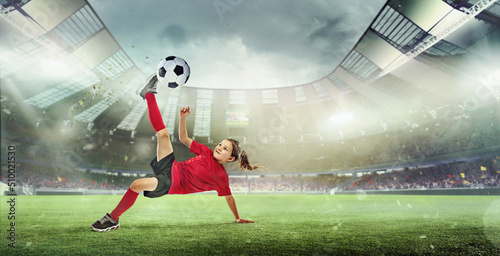 Portrait of little girl, football player in red uniform hitting ball and falling isolated over open air stadium. © Lustre