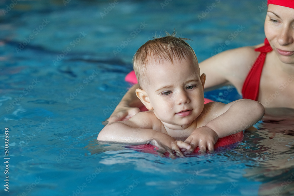 Small baby boy learns to swim on swimming board in pool with support of mother.