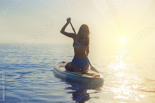 Asian beautiful girl in a blue bikini is engaged in water sports. She sits with her back on a surfboard with a paddle in the ocean against the background of a sunset. Woman on paddleboarding , Sup. © Tetiana