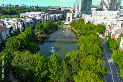 an aerial shot of the silky green waters of the lake with a rust colored metal bridge over the water surrounded by lush green trees, grass and plants with office buildings and apartments photo