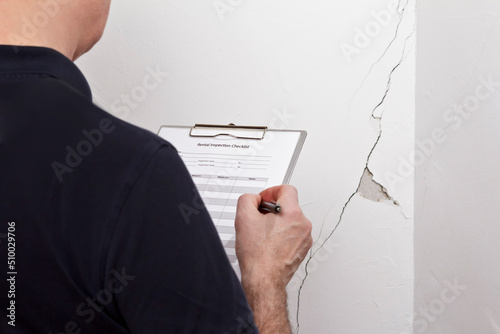 Murais de parede Man with inspection checklist in front of a white wall with a long crack or rip and a piece of plaster missing, rental damage concept