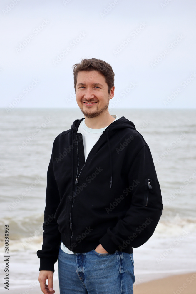 A young man in a black hoodie on the beach against the backdrop of the sea. Brown hair, blue eyes, smiling A young man in a black hoodie on the beach against the backdrop of the sea. Brown hair, blue eyes, smiling