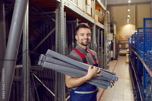 Warehouse worker at building materials store stands with plumbing pipes for sewage in one of aisles. Portrait of smiling young man in coveralls holding gray plastic pipes and looking at camera.