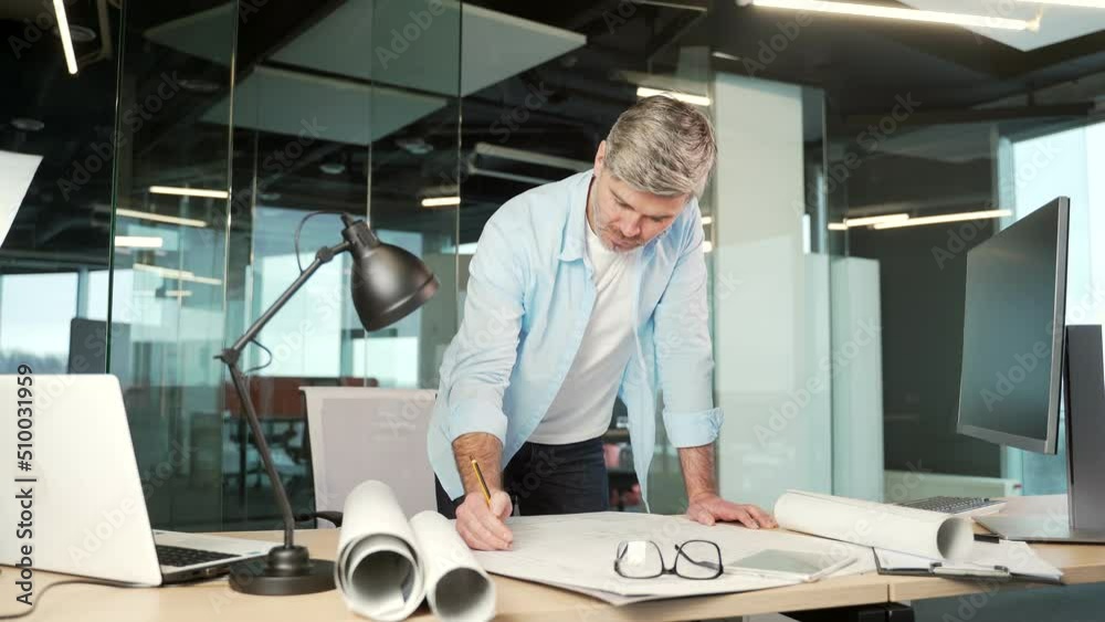 mature creative designer, architect working in the office at work a computer desk with drawings. Handsome senior man doing project work standing at workplace indoor sketching a housing plan building mature creative designer, architect working in the office at work a computer desk with drawings. Handsome senior man doing project work standing at workplace indoor sketching a housing plan building