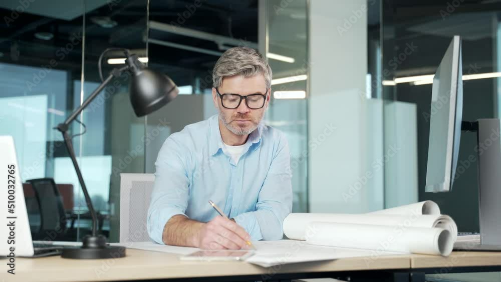Experienced mature creative designer, architect working in the office at computer desk with drawings. Handsome senior man doing project work sitting at workplace sketching a housing plan building
