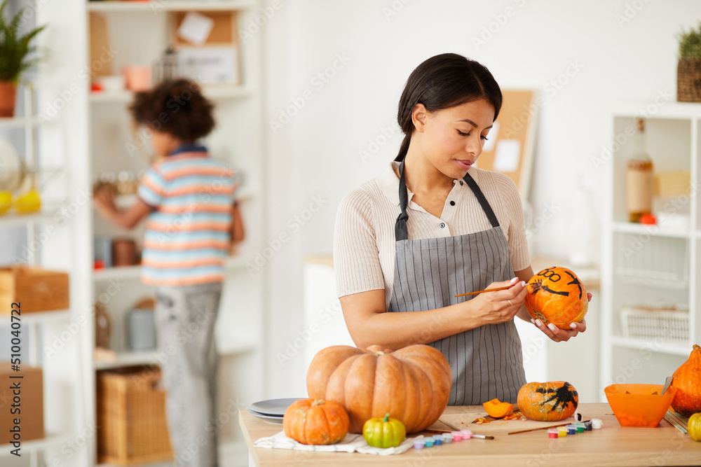 Content attractive young woman in gray apron standing at kitchen counter and making Halloween drawings on pumpkins