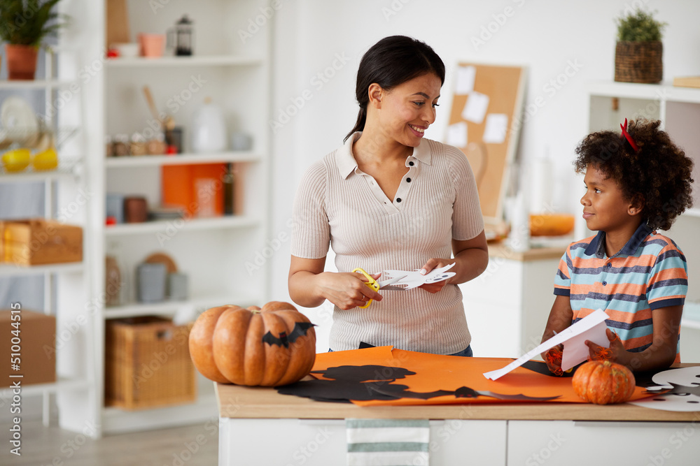 Positive young black woman cutting out paper ghost with son, she enjoying handmade process with son