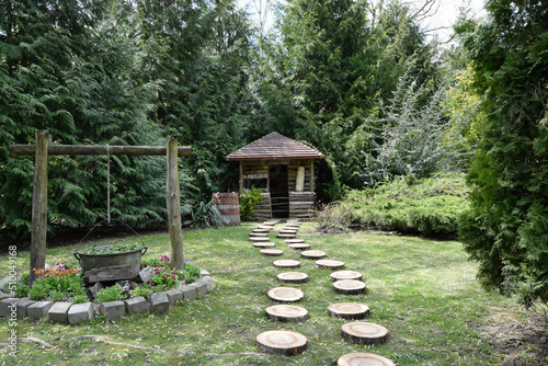 Beautiful view of a small wooden house surrounded by green trees. Path of round pieces that wood taking the place of the alley. Garden decorations