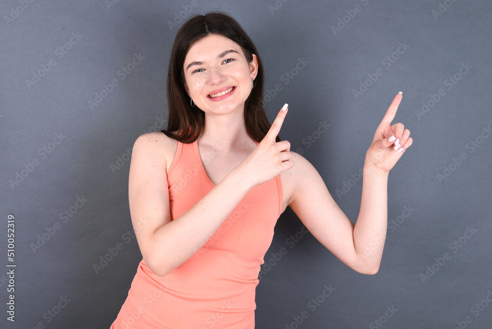 Caucasian woman wearing orange T-shirt over grey wall indicating finger empty space showing best low prices, looking at the camera Caucasian woman wearing orange T-shirt over grey wall indicating finger empty space showing best low prices, looking at the camera