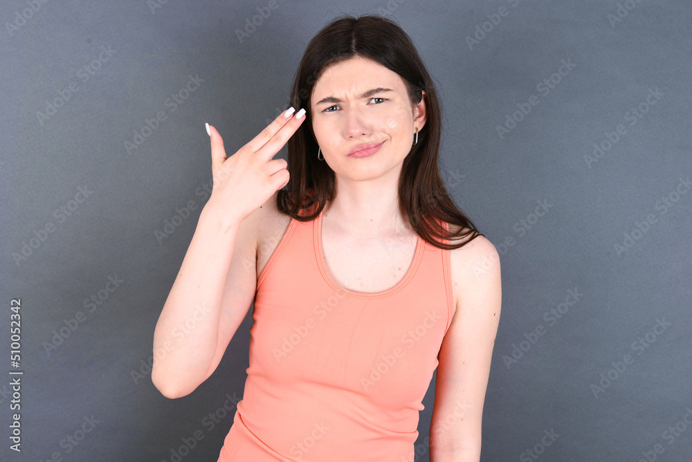 Unhappy Caucasian woman wearing orange T-shirt over grey wall makes suicide gesture and imitates gun with hand, curves lips, keeps two fingers on temple Unhappy Caucasian woman wearing orange T-shirt over grey wall makes suicide gesture and imitates gun with hand, curves lips, keeps two fingers on temple
