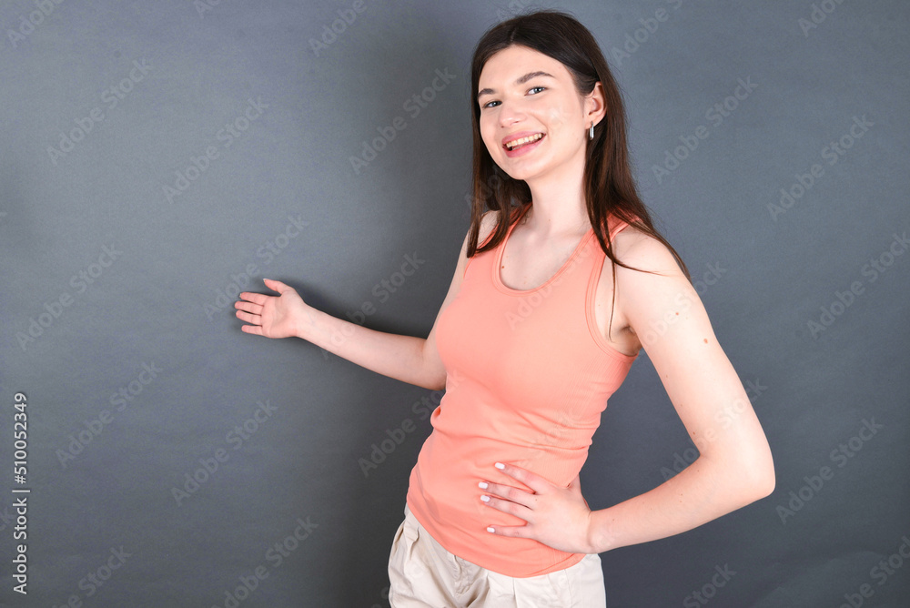 Caucasian woman wearing orange T-shirt over grey wall feeling happy and cheerful, smiling and welcoming you, inviting you in with a friendly gesture Caucasian woman wearing orange T-shirt over grey wall feeling happy and cheerful, smiling and welcoming you, inviting you in with a friendly gesture
