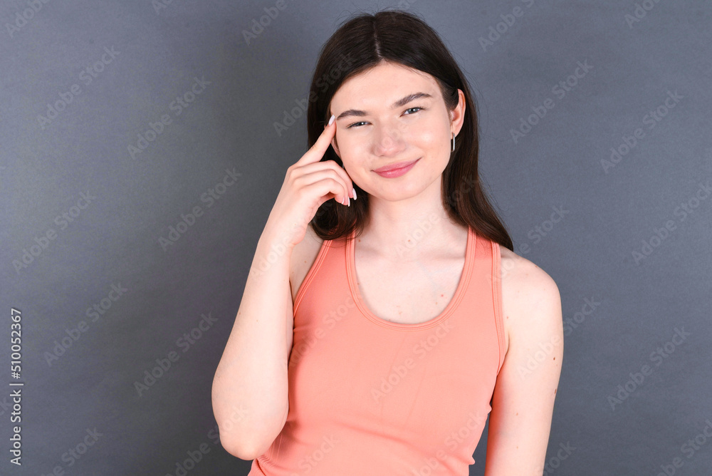 Caucasian woman wearing orange T-shirt over grey wall tries to memorize something, keeps fore finger on temple, reminds information for exam,