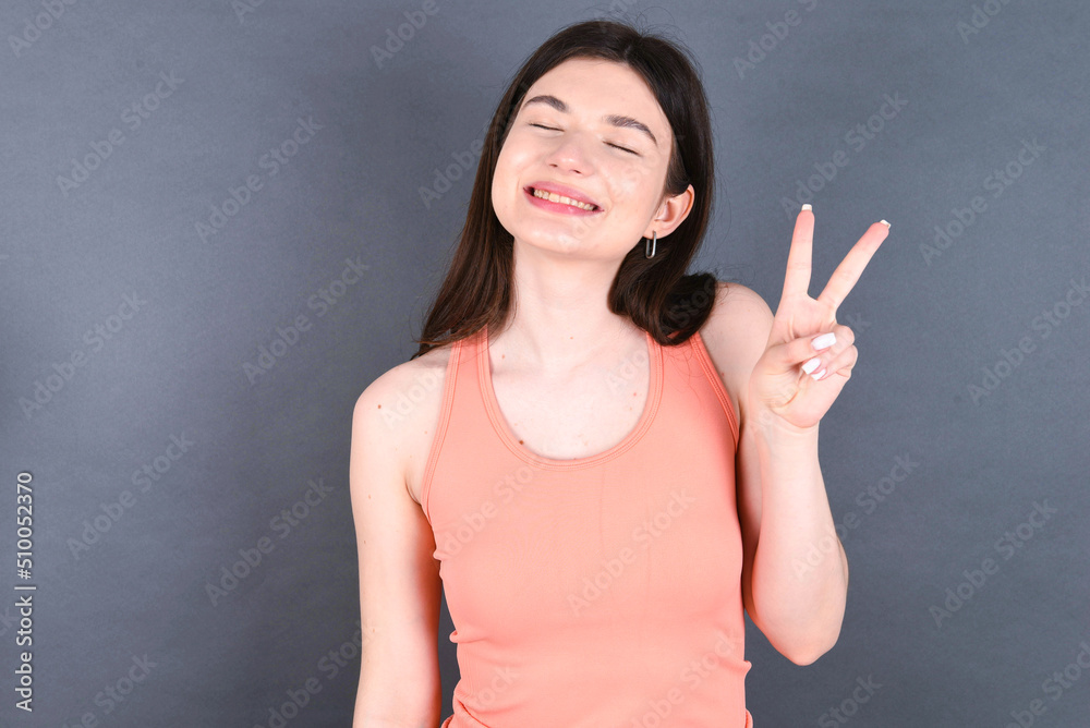 Caucasian woman wearing orange T-shirt over grey wall smiling with happy face winking at the camera doing victory sign. Number two. Caucasian woman wearing orange T-shirt over grey wall smiling with happy face winking at the camera doing victory sign. Number two.