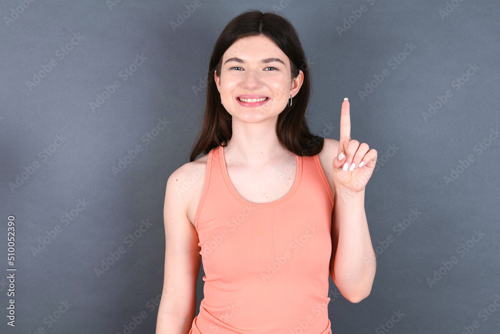 Caucasian woman wearing orange T-shirt over grey wall showing and pointing up with finger number one while smiling confident and happy. Caucasian woman wearing orange T-shirt over grey wall showing and pointing up with finger number one while smiling confident and happy.
