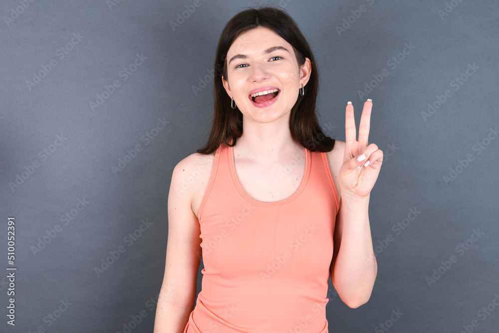 Caucasian woman wearing orange T-shirt over grey wall showing and pointing up with fingers number two while smiling confident and happy. Caucasian woman wearing orange T-shirt over grey wall showing and pointing up with fingers number two while smiling confident and happy.