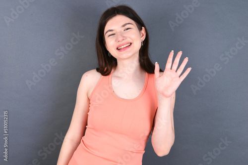Caucasian woman wearing orange T-shirt over grey wall Waiving saying hello happy and smiling, friendly welcome gesture.