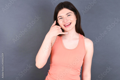 Caucasian woman wearing orange T-shirt over grey wall smiling doing phone gesture with hand and fingers like talking on the telephone. Communicating concepts.