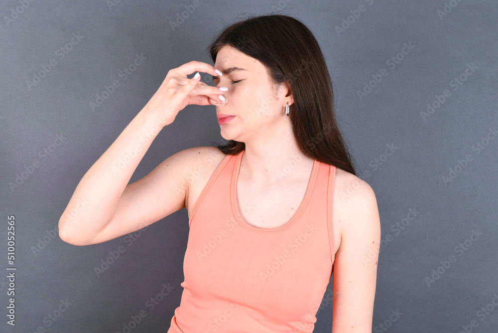 Caucasian woman wearing orange T-shirt over grey wall smelling something stinky and disgusting, intolerable smell, holding breath with fingers on nose. Bad smell Caucasian woman wearing orange T-shirt over grey wall smelling something stinky and disgusting, intolerable smell, holding breath with fingers on nose. Bad smell
