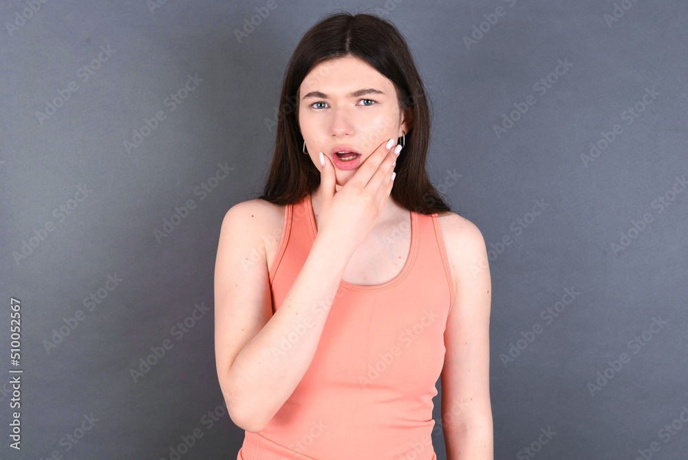 Caucasian woman wearing orange T-shirt over grey wall Looking fascinated with disbelief, surprise and amazed expression with hands on chin Caucasian woman wearing orange T-shirt over grey wall Looking fascinated with disbelief, surprise and amazed expression with hands on chin
