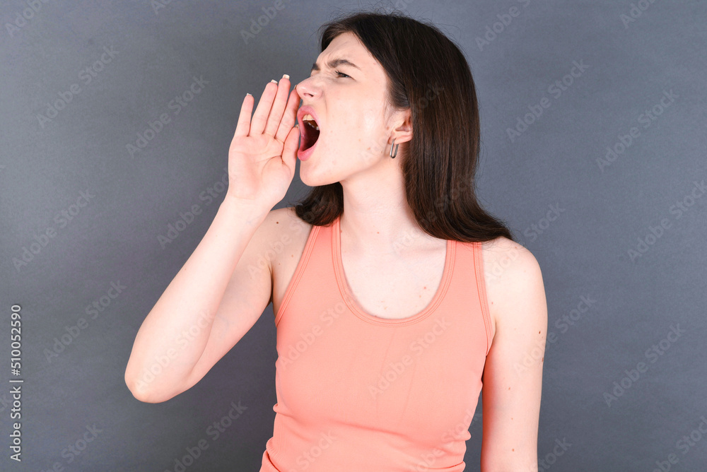 Caucasian woman wearing orange T-shirt over grey wall shouting and screaming loud to side with hand on mouth. Communication concept. Caucasian woman wearing orange T-shirt over grey wall shouting and screaming loud to side with hand on mouth. Communication concept.